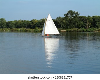 Yacht sailing on the Norfolk broads. Full white sail calm water trees in the background