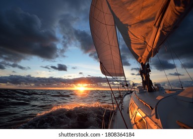 Yacht sailing in an open sea at sunset. Close-up view of the deck, mast and sails. Clear sky after the rain, dramatic glowing clouds, golden sunlight, waves and water splashes, cyclone. Epic seascape