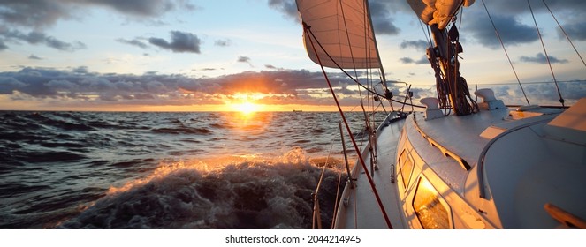 Yacht, die bei Sonnenuntergang im offenen Meer segelt. Nahaufnahme von Deck, Mast und Segel. Klare Himmel nach dem Regen, dramatische leuchtende Wolken, goldenes Sonnenlicht, Wellen und Wasser splash, Zyklon. Epische Jahreszeit