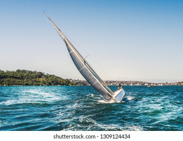 A Yacht Tilting, Heeling In The Water It Has A Large Silver Grey Sail And Is Cutting Through The Blue Water Of Sydney Harbour, Australia On A Summer Day. An Unrecognisable Man Is Sailing The Boat.