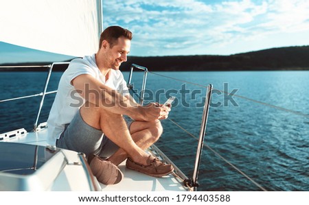 Yachting Concept. Man Using Smartphone App Sitting On Deck Relaxing Sailing Across The Sea Outdoors. Copy Space