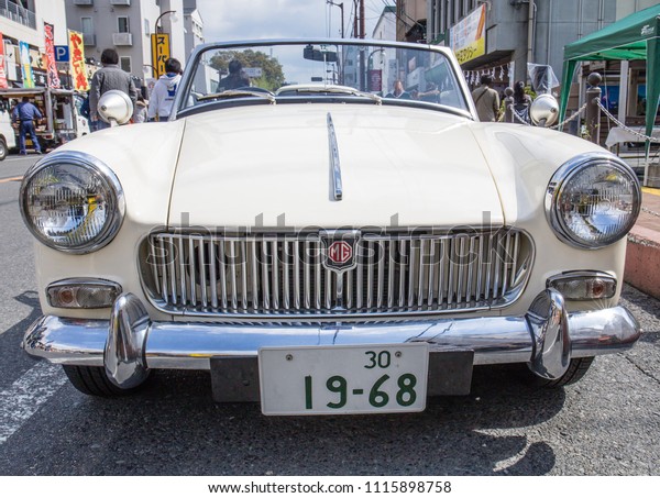YAMAGUCHI, JAPAN - APRIL 8, 2018: A front\
view close up of the shiny chrome bumper, grill, and hood of a\
vintage, cream white MGB roadster sports\
car.