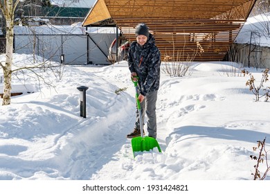 The Yard Was Covered With Snow After A Heavy Snowfall. A Man Cleans The Garden Paths From Snow On A Sunny Day In Winter. The Gardener Takes Care Of The Garden And Clears The Snowdrifts With A Shovel.