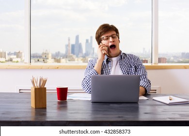 Yawning European Man Having Mobile Phone Conversation At Office Desk With Laptop, Business Report, Coffee Cup And Stationery Items. Front View