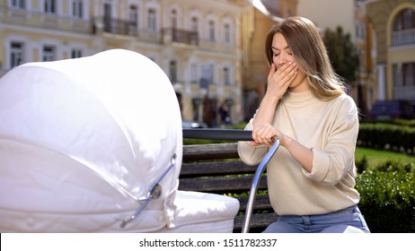 Yawning mother swinging infant carriage sitting on park bench, lack of sleep