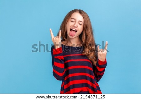 Yeah, awesome ! Portrait of excited rocker woman in sweater showing rock and roll hand gesture, crazy devil horns with fingers and yelling. Indoor studio shot isolated on blue background.