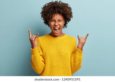 Yeah, feeling cool and awesome. Excited happy young African American woman lifts mood from loud music, shows rock n roll gesture, glad to visit concert of favourite band, exclaims loudly with joy.