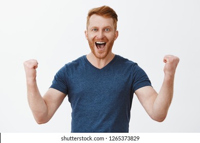 Yeah, good work man. Happy cheerful good-looking redhead strong man, raising clenched fists in hooray gesture, smiling broadly, triumphing from successful deal or great news, being joyful and thrilled