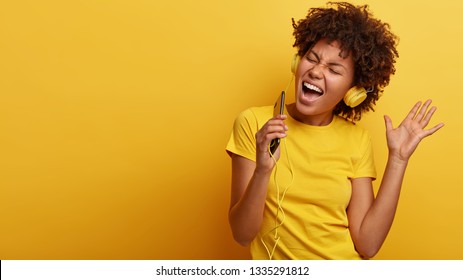 Yeah, I am pleased. Joyful lovely dark skinned woman raises hand in dance move, likes great sound in new headphones, sings loudly, carries cellular, smiles broadly, wears yellow casual clothes