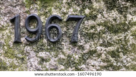 The year 1967 cast in metal and affixed to a stone – a detail of an inscription produced that year. The stone is covered with lichen