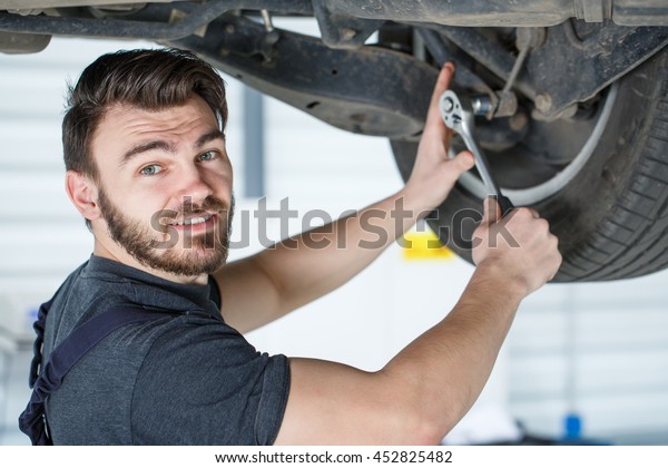 Years of experience. Closeup portrait of a\
smiling mechanic working on a car undercarriage with a wrench\
smiling to the camera over his shoulder\
