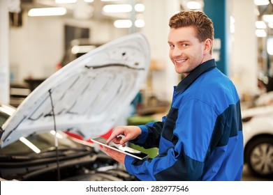 Years Of Mechanical Experience. Confident Young Man Working On Digital Tablet And Smiling While Standing In Workshop With Car In The Background