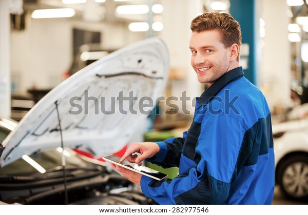 Years of mechanical experience. Confident\
young man working on digital tablet and smiling while standing in\
workshop with car in the\
background