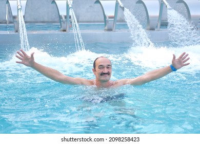 Yekaterinburg, Russia - August 16, 2021: Smiling mature asian man looking at camera opening your hands in outdoor thermal pool with hydromassage over background of injectors with water jets.