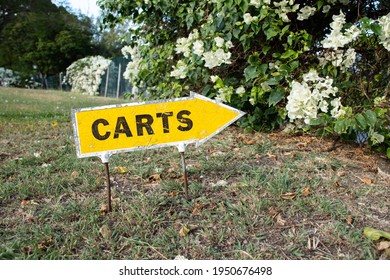 A Yellow Arrow-shaped Sign For Golf Carts With A White Border, Attached To Rusted Stakes Poking Into The Grass In Front Of A Cluster Of White Bougainvillea On A Private Golf Course In Barbados. 