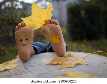 Yellow Autumn Leaf Between The Toes On The Bare Feet Of A Toddler Child. Painted Smile On The Feet. Indulge, Positive Thinking, Happy Childhood. Hello, Autumn. Seasonal Fun Photo Ideas