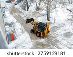 Yellow backhoe loader clearing heavy, slushy snow and ice from a city street during the winter season.