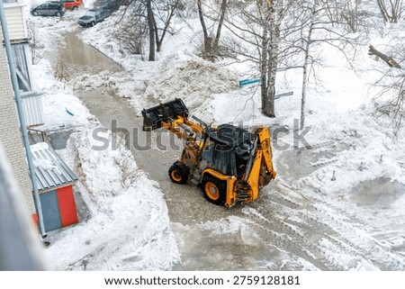 Yellow backhoe loader clearing heavy, slushy snow and ice from a city street during the winter season.