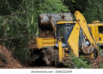 Yellow backhoe loader clearing mud and soil with bucket on rural road. Powerful construction machinery digging dirt and debris in lush green forest for site cleanup