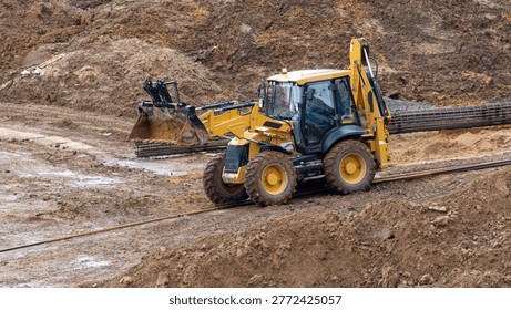 Yellow backhoe loader digs into muddy terrain. Its large tires sink into wet earth. The machine moves soil for land development. Construction site activity is clearly shown