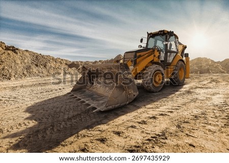 A yellow backhoe loader digs into the earth on a construction site as the sun sets, casting long shadows on the rugged ground.