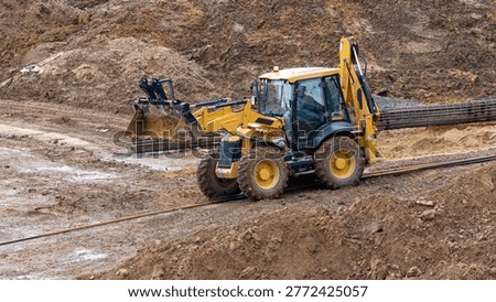 Yellow backhoe loader digs into muddy terrain. Its large tires sink into wet earth. The machine moves soil for land development. Construction site activity is clearly shown