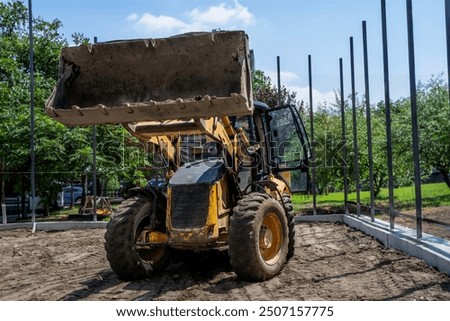 A yellow backhoe loader stands idle at a construction site surrounded by trees with skyscrapers visible in the background. The machinery's worn appearance suggests it has been in use for some time.