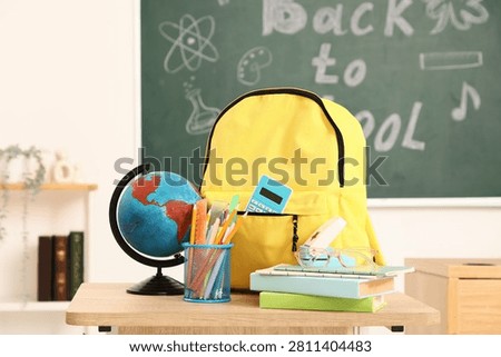 Yellow backpack with school supplies, globe, and books on a wooden desk in a classroom setting