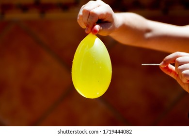 Yellow Balloon Filled With Water In A Child's Hand And A Needle In The Other Hand