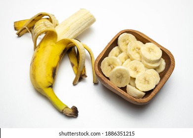 yellow and bitten banana lies on a white background, next to a wooden cup with sliced bananas, on the table