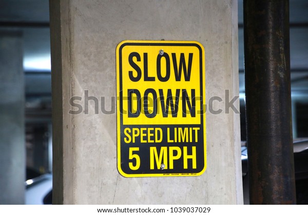 Yellow and Black Slow\
Down Speed Limit 5 Mph Sign in a Parking Garage with Cars Out of\
Focus in the Background