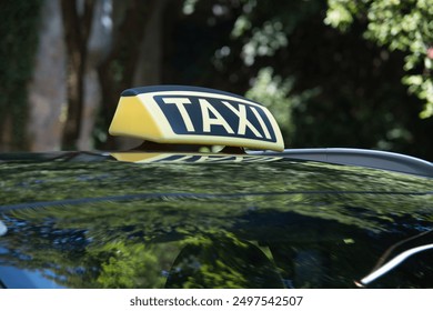 A yellow and black taxi sign is on the roof of a car. The sign is in the shade of a tree - Powered by Shutterstock