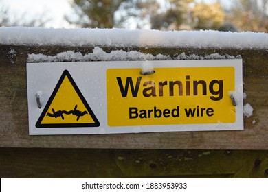 Yellow, Black And White Rectangular Warning Barbed Wire Sign With Barbed Wire Icon Inside Triangle. On Wooden Fence With Snow On Top Of Fence. Blurred Background.