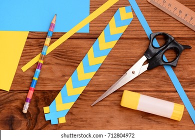 Yellow Blue Paper Bookmark On Wood Background. Scissors, Glue Stick, Colored Paper Sheets, Ruler, Pencil On Wooden Table. Light Art Project. Ukrainian Paper Craft For Kids With Folding Paper. Top View