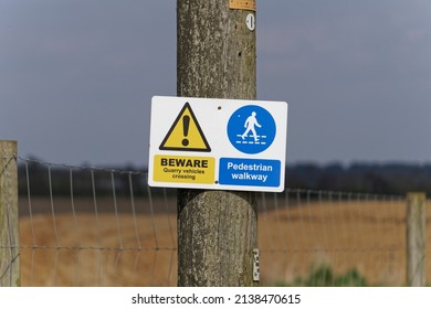A yellow and blue warning sign with the words Beware quarry vehicles crossing Pedestrian Walkway on the perimeter of a quarry in the English countryside
