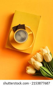 Yellow book on yellow background and coffee in yellow cup on top of the book. with white tulips and chocolate chips