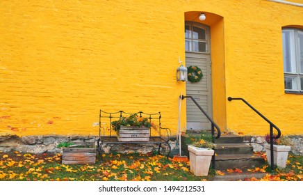 Yellow Brick House Entrance With Seasonal Wreath On Door And Porch Window On Autumn Day With Fall Leaves On The Ground. Iron Bench Furniture And Wooden Box Garden Flowers - Vintage Autumn Decoration