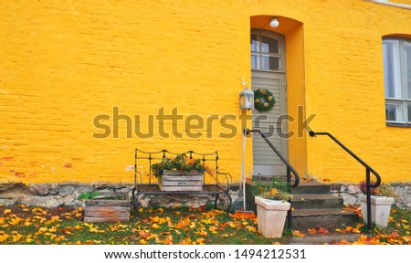 Yellow brick house entrance with seasonal wreath on door and porch window on autumn day with fall leaves on the ground. Iron bench furniture and wooden box garden flowers - vintage autumn decoration