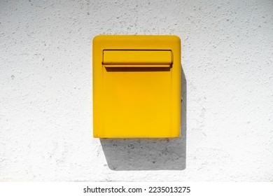 Yellow bright metal mailbox hangs on white rough wall casting shadow from sun. Stylish detail in interior of house authentic street box for letters postcards. Background good news holiday messages