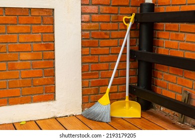 Yellow Broom And Dustpan Leaning Against The Red Brick Wall At The Corner Of A House Patio.