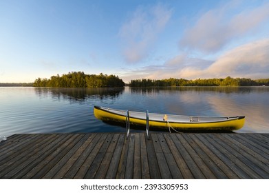 A yellow canoe is securely fastened to a rustic wooden dock nestled in the serene beauty of Muskoka, Ontario, Canada. In the distance, quaint cottages dot the landscape across the tranquil lake. - Powered by Shutterstock