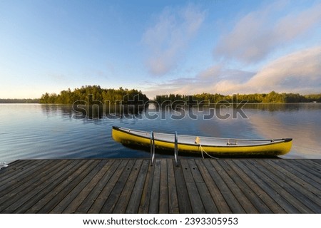 A yellow canoe is securely fastened to a rustic wooden dock nestled in the serene beauty of Muskoka, Ontario, Canada. In the distance, quaint cottages dot the landscape across the tranquil lake.
