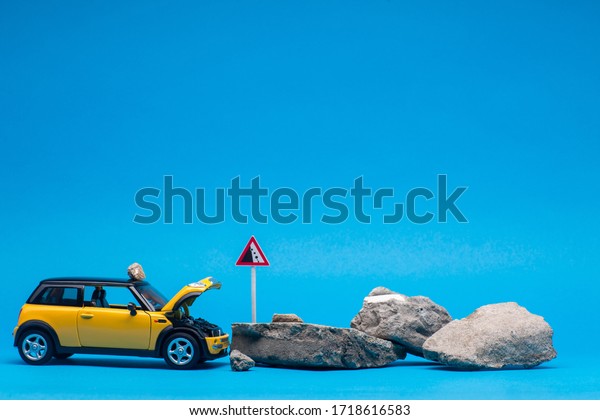 An yellow car with its hood open next to a\
bunch of stones, on blue\
background.