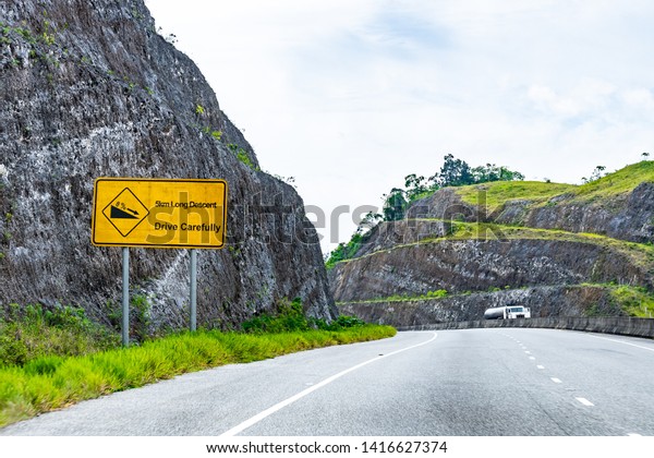 Yellow Caution 5km Long Descent downhill 8% decline\
street traffic arrow symbol sign on dual carriageway highway\
through scenic mountain countryside where cars drive on left hand\
side of the road.
