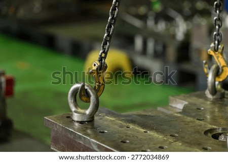 Yellow chain hook locks into steel lifting eye above heavy die block, supporting controlled hoist connection, safe mold transfer, and maintenance preparation in modern manufacturing workshop