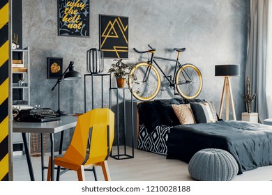 Yellow Chair At Desk In Grey Bedroom Interior With Posters And Bicycle Above Black Bed. Real Photo