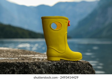 A yellow children's rubber boot (gumboot) stands on a stone on the shore of a lake against a blurred background of mountains and water. - Powered by Shutterstock