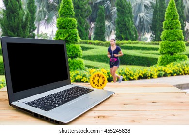 Yellow clock and computer are on the table, blur image of woman jogging in the morning as background.