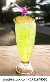 Yellow Colored Tropical Drink Made From Pineapple Juice, And Served Over Ice In A Tall Glass, Garnish With Pineapple Wedge And Pink Spiral Straw. Blur Backyard Background.