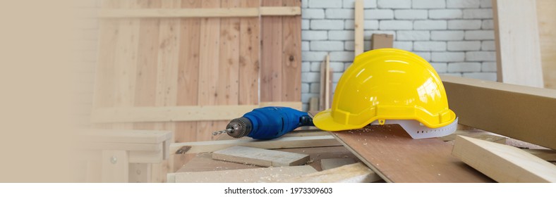 The Yellow Construction Helmet And The Blue Electric Drill Were Left Aside During Lunch Time Brake. A Desk Full Of Hand Tools And Wood Piles. Morning Work Atmosphere In The Workshop Room.